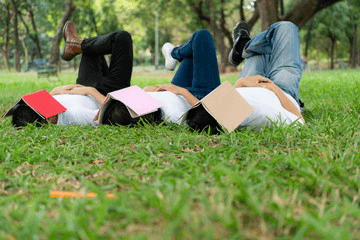 Three students lying in the grass sleeping with their textbooks covering their faces and their legs crossed